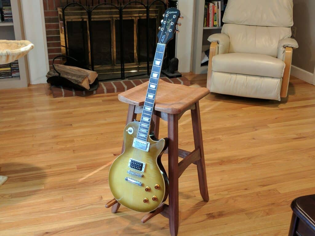 A modified version of the bench stool project with two prongs mounted to one of the bottom rails so that the stool can double as a guitar stand when not in use. The seat and guitar rests are mahogany, and the frame is sapele. (The pictured guitar belongs to my brother. I did not make it.)