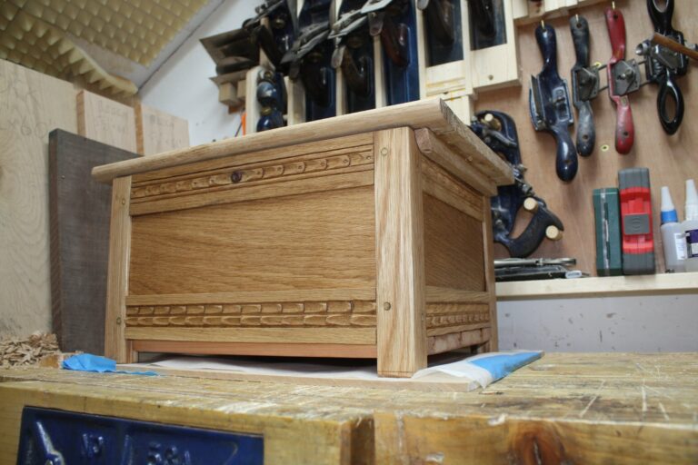 Faux-jacobian frame-and-panel blanket chest in oak sized for baby blankets as a gift for a coworker who'd just had his first child. Drawbored mortise and tenon joints, low relief carving, tongue-and-grooved cedar base, finished with Osmo wood wax and beeswax.
